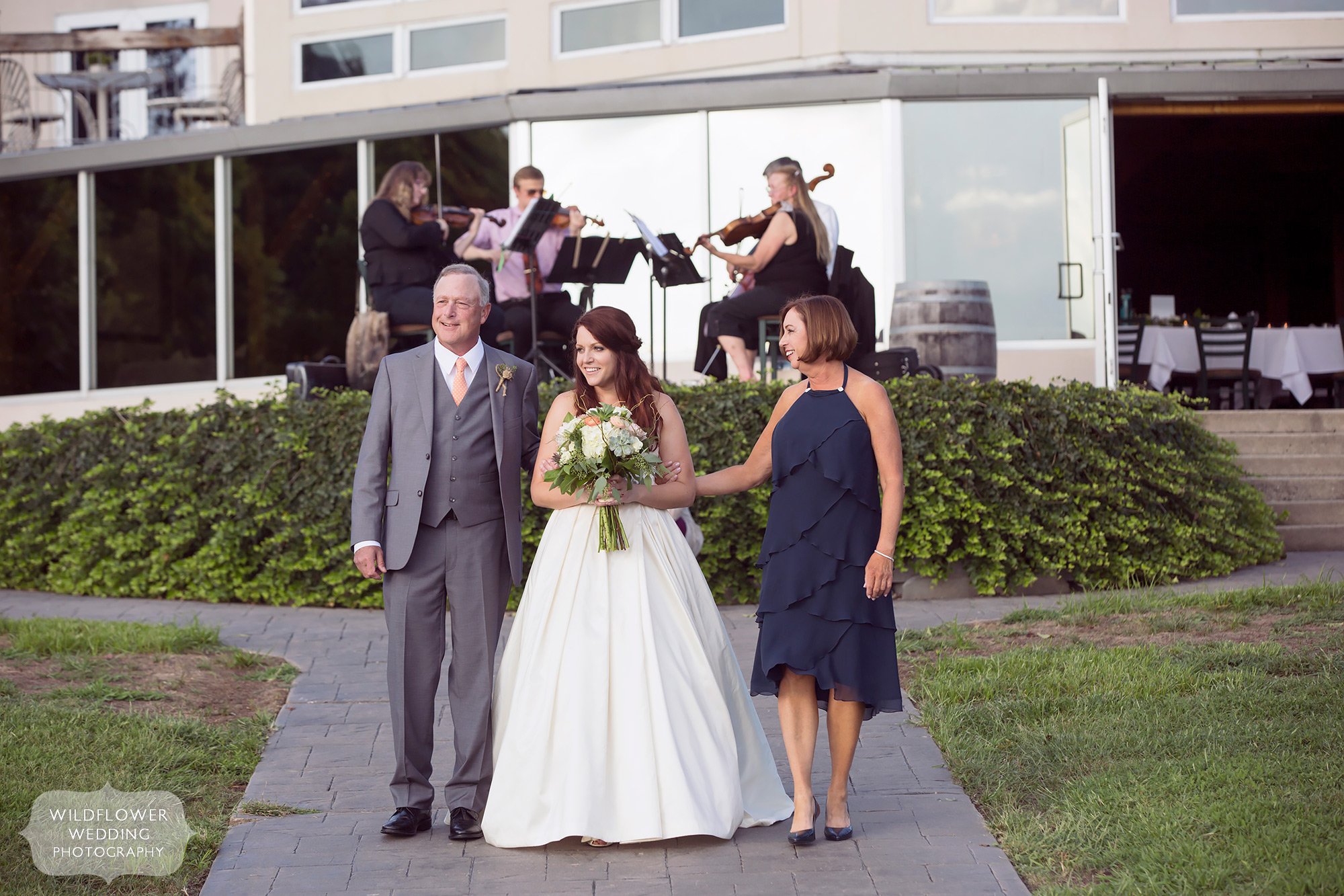 Bride walks with her parents down the aisle during her summer outdoor wedding at Les Bourgeois in MO.