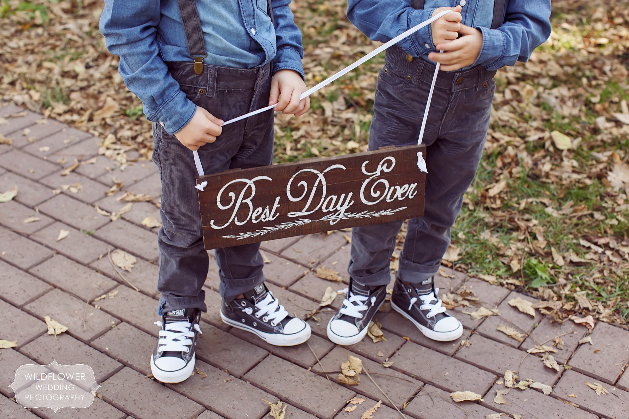 Twin ring bearers wearing converse shoes hold a wooden wedding sign with script font that reads, "Best Day Ever".