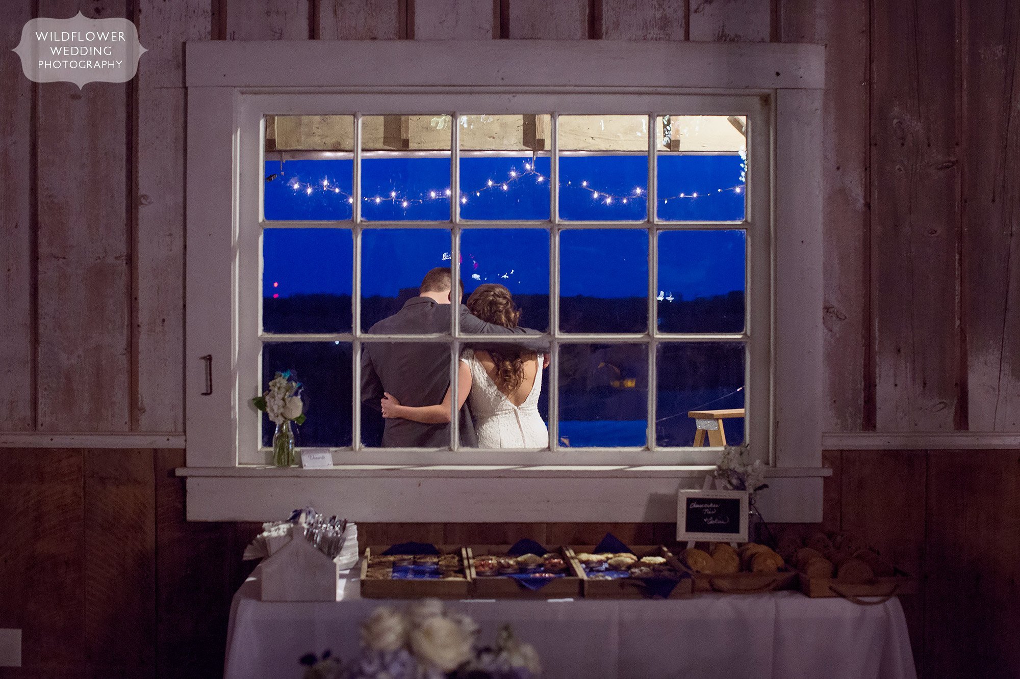 Twilight photo of the bride and groom on the porch outside of a window.