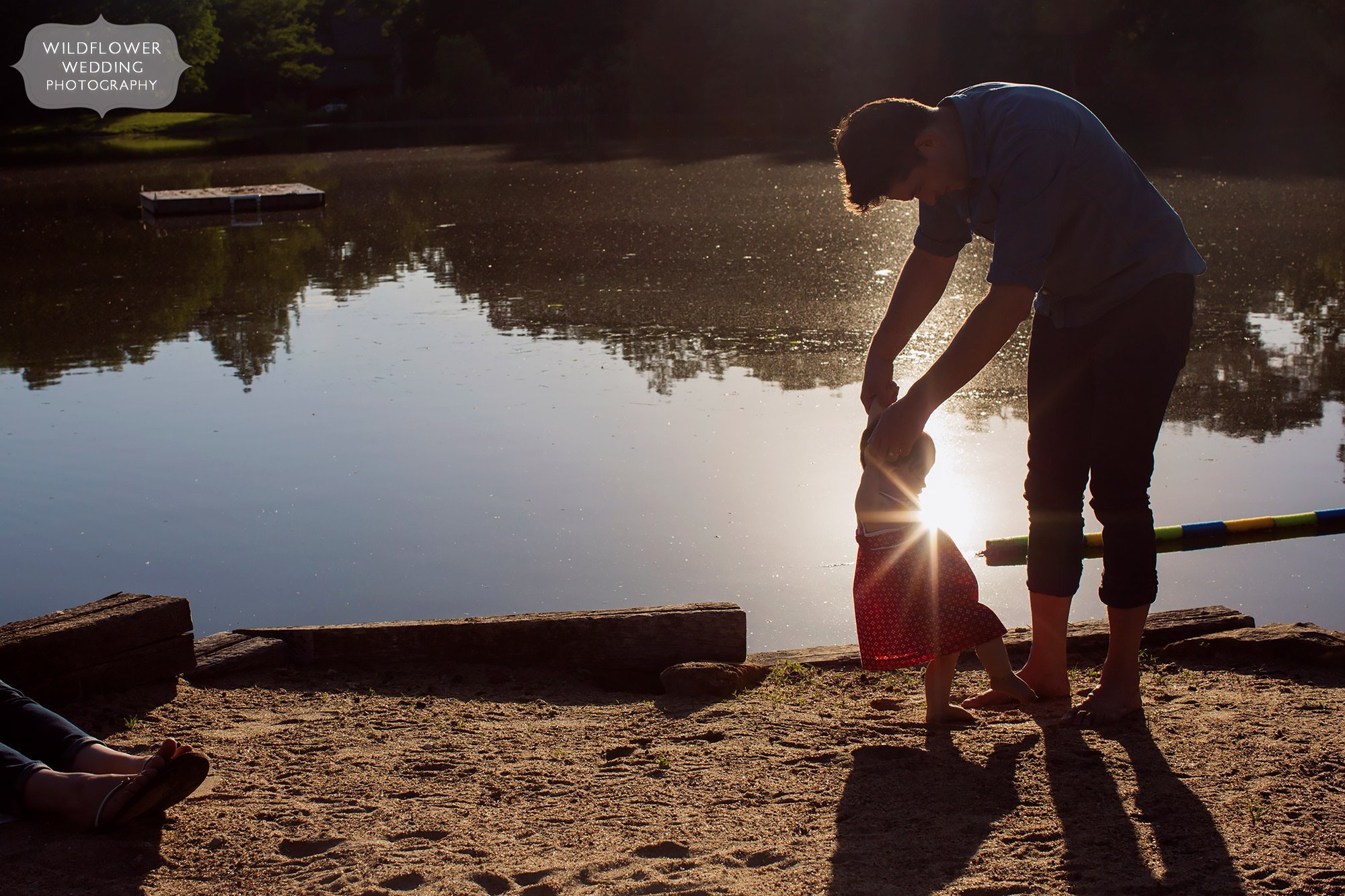 Artsy photo of a dad helping his baby walk at dusk with a sunbursty sunset light.