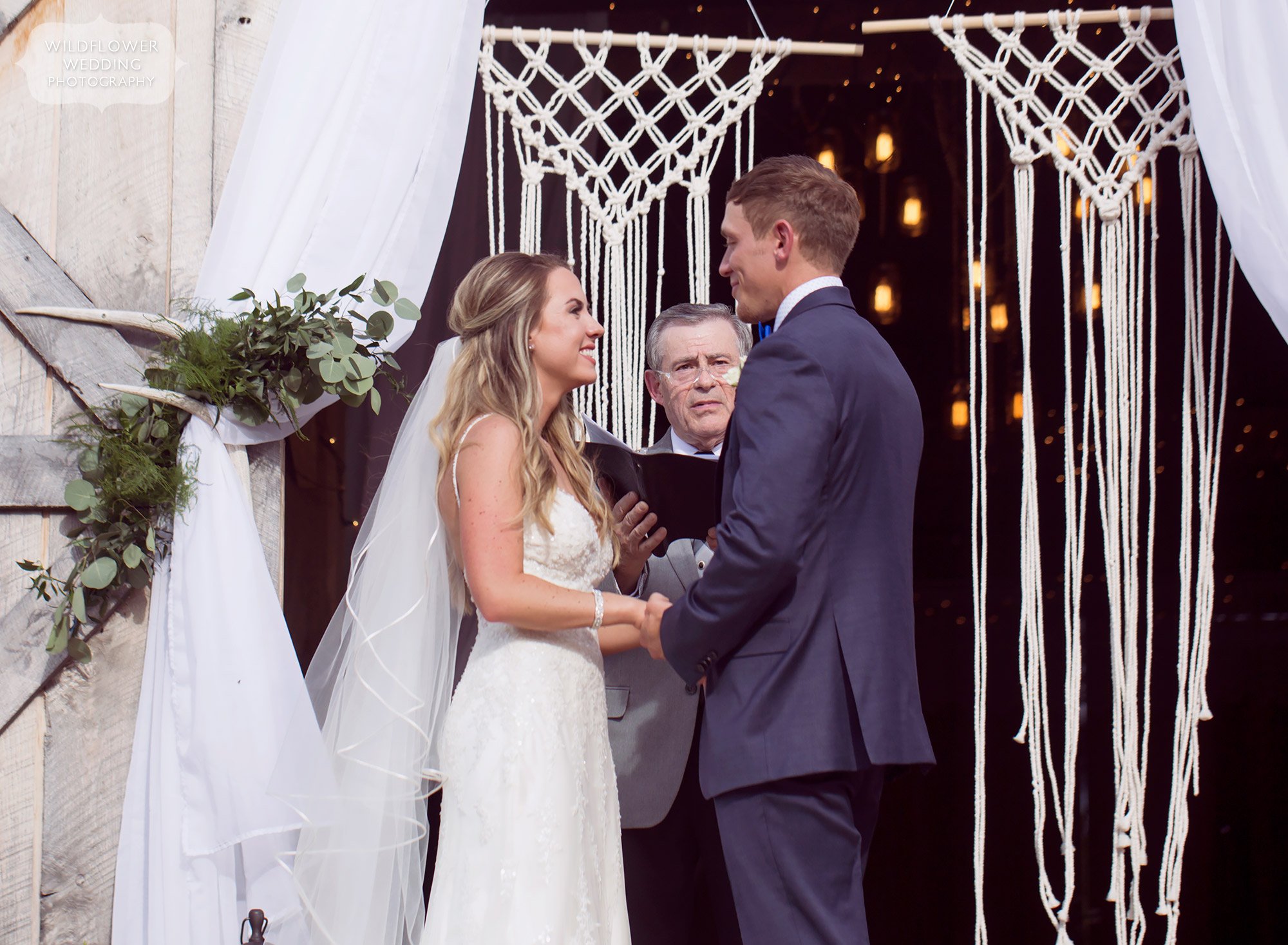 The bride and groom exchange vows in front of this white woven macrame backdrop in the barn doorway at Kempker's Back 40.