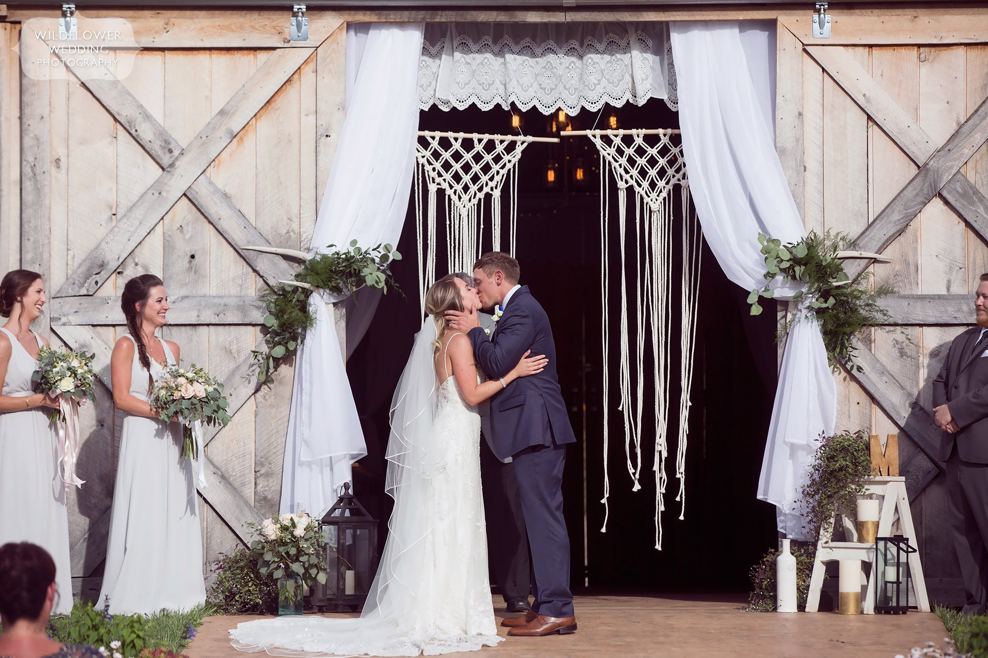 The bride and groom kiss in front of the crocheted macrame backdrop in the barn at Kempker's Back 40.