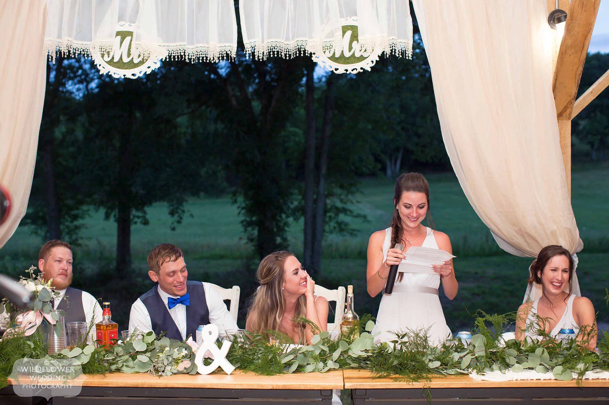 The maid of honor gives a speech while the bride and groom laugh under the covered pavilion for dinner.