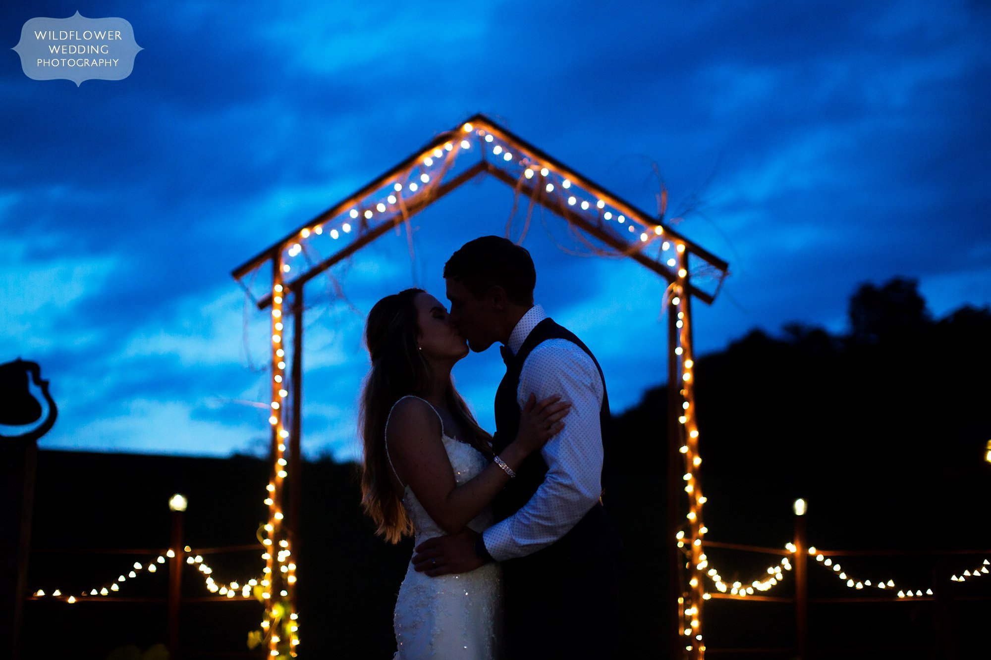 Bride and groom kiss under a twinkle light arch during twilight blue light sky in southern MO.