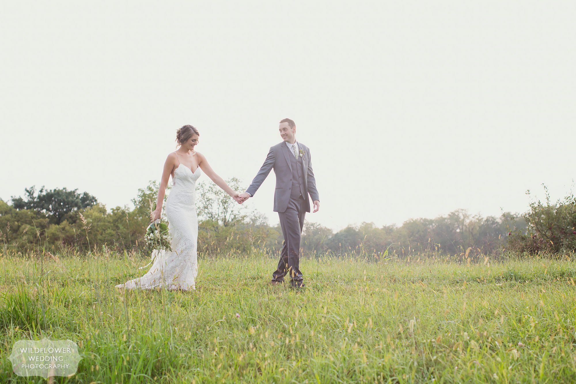Soft light wedding photography of bride and groom walking through field holding hands in Weston, MO.