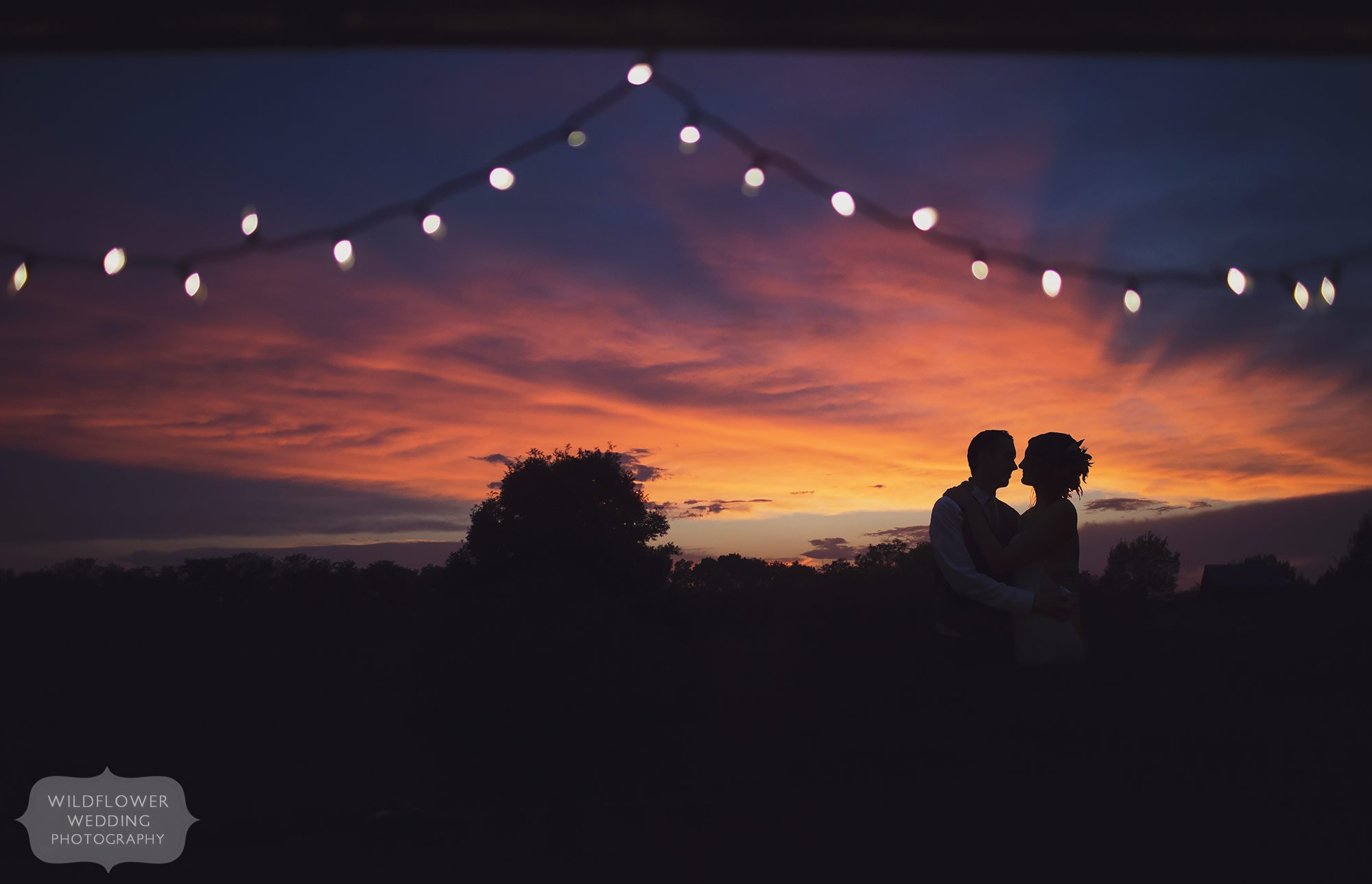 Amazing wedding photo of the bride and groom silhouetted with colorful sunset behind them and twinkle lights in Weston, MO.