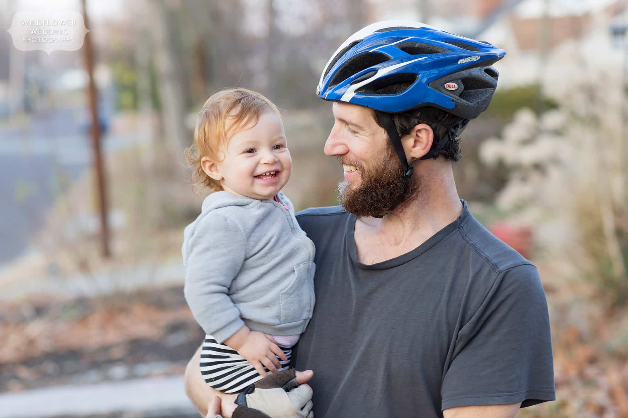 Dad wearing a bicycle helmet poses with his 18 month old toddler outside. 