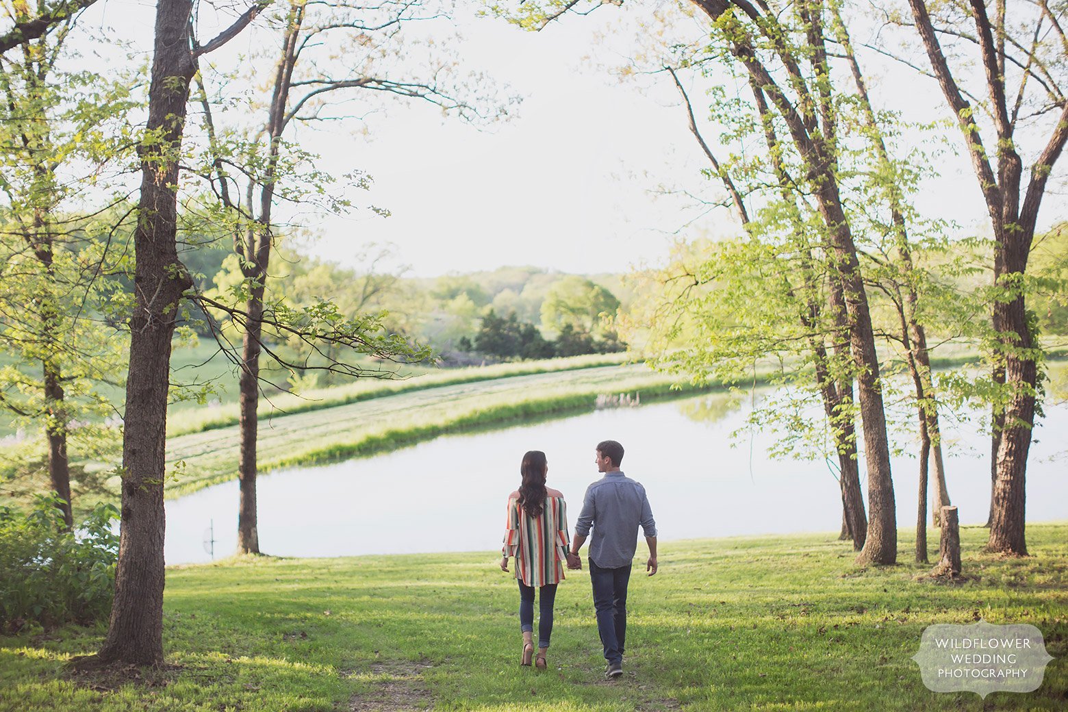 Couple holds hands while walking toward pond during a rural Missouri engagement photo session.