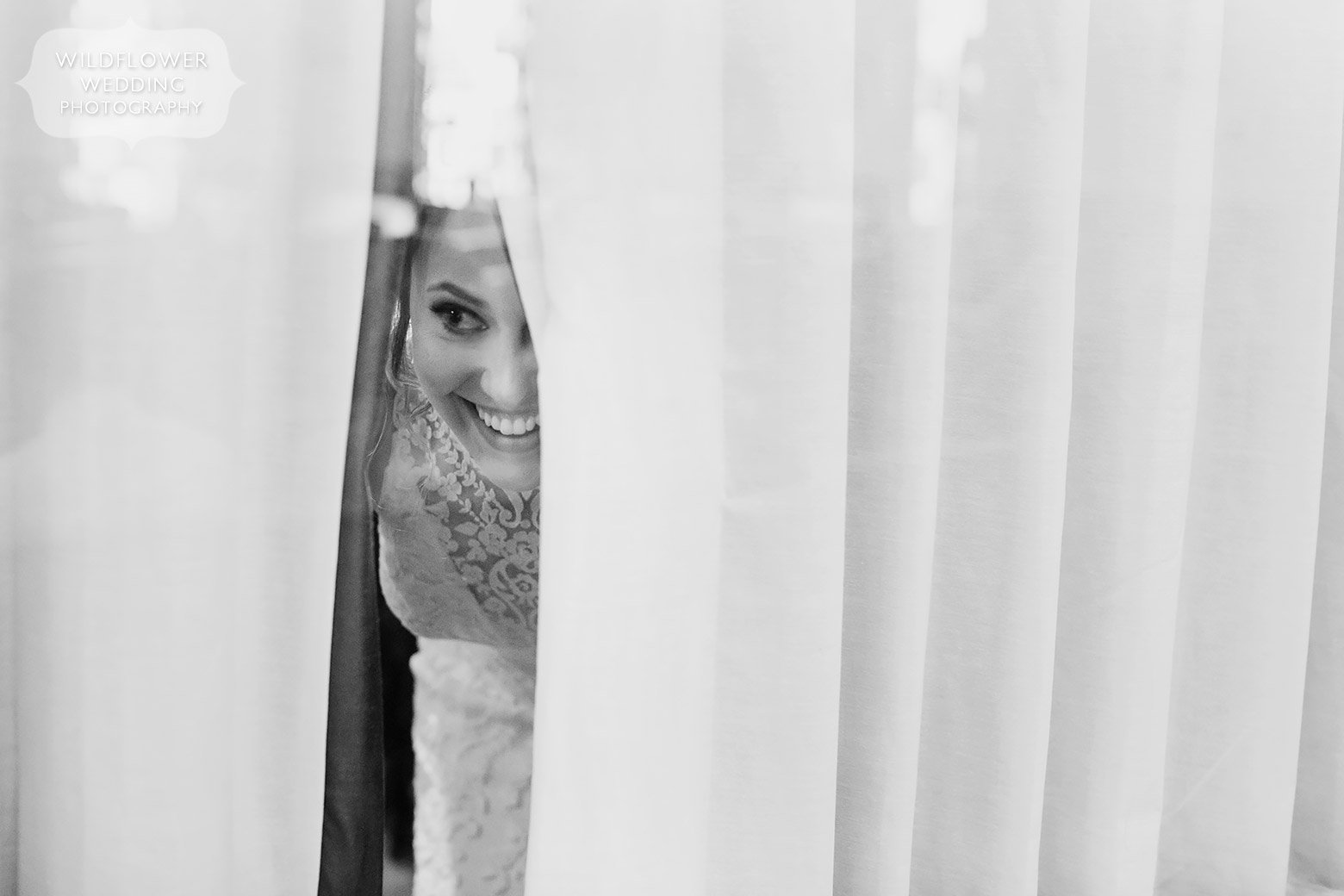 Candid wedding photography of bride looking out of curtains at guests.