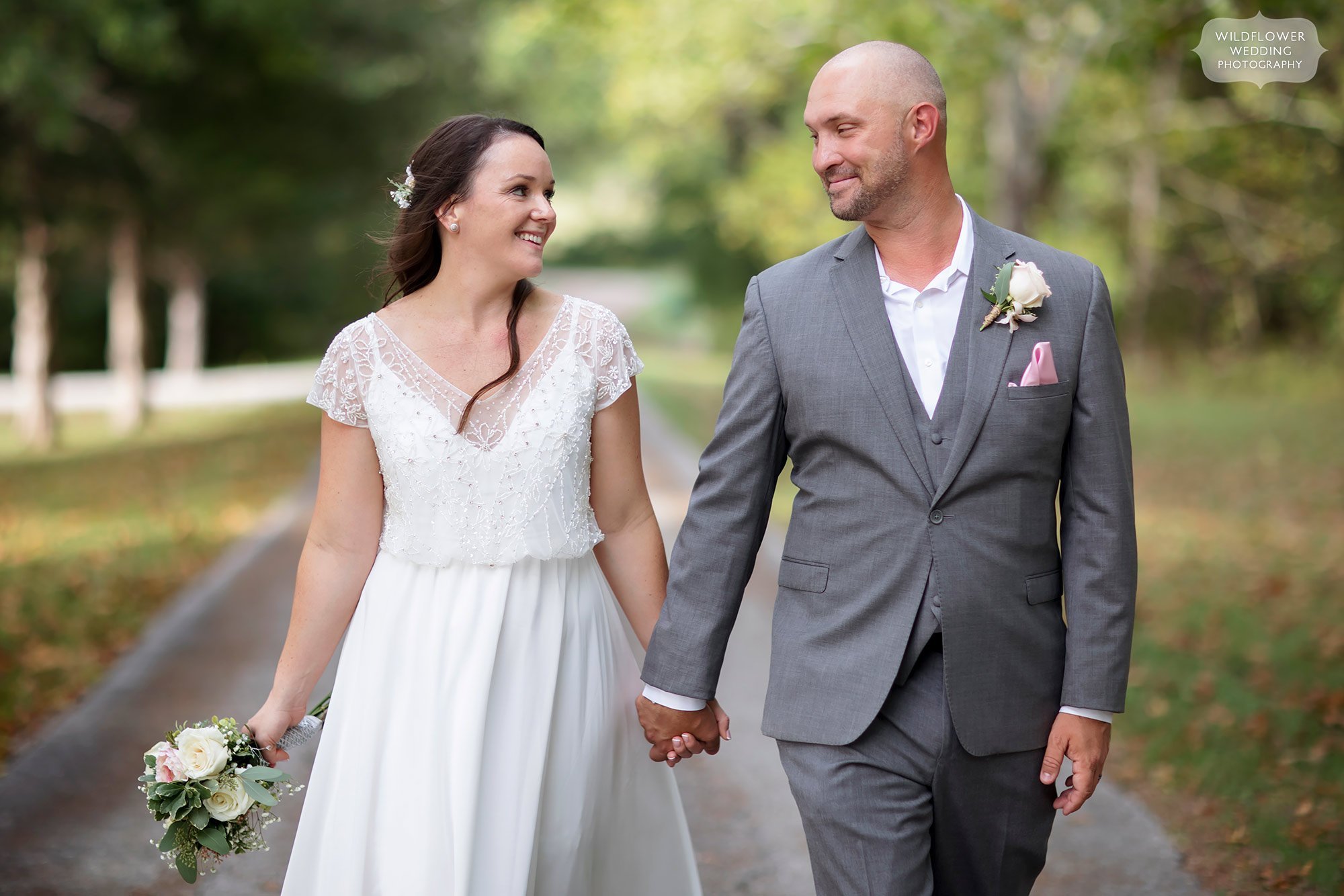 Bride and groom hold hands and walk down gravel road.