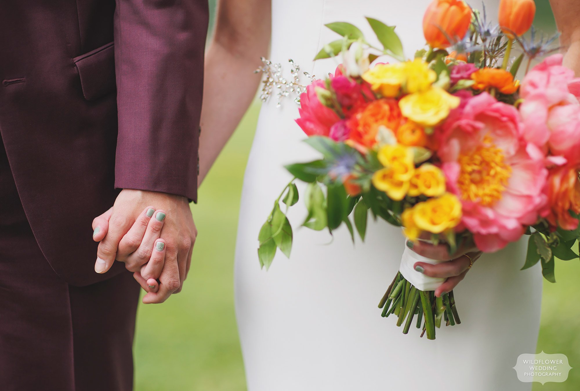 Bride and groom hold hands with blue nail polish and colorful wedding bouquet.